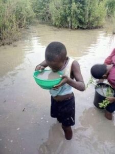 Photo : un enfant noir boit de l'eau marron dans une bassine verte. Il a les pieds dans l'eau d'une rivière marron aussi.